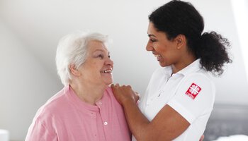Eine Pflegerin der Caritas legt die Hand auf die Schulter einer Seniorin und die beiden lachen sich gegenseitig an | © Picasa