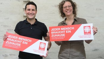 Mann und Frau halten jeweils ein "Jeder Mensch braucht ein Zuhause"-Schild | © Caritas München Oberbayern
